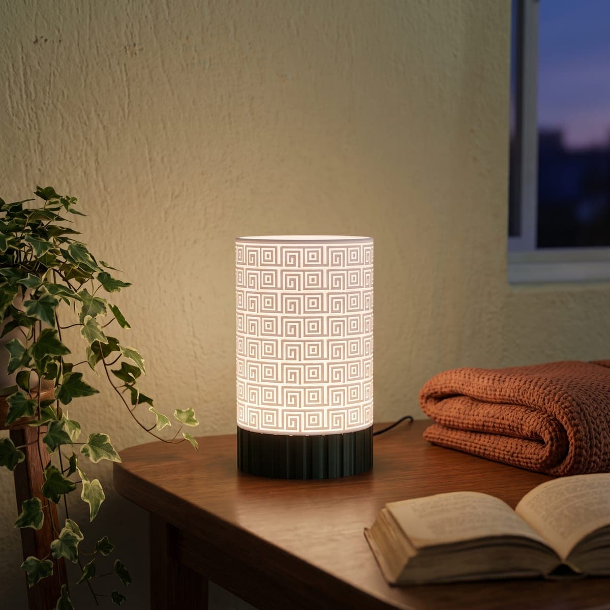 Lamp with a white Greek key shade on a wooden bedside table, beside a plant, a folded blanket and an open book